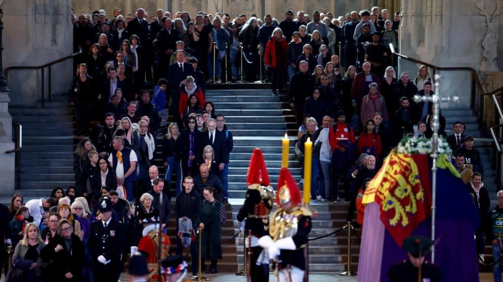 A large group of people line either side of a staircase. There is a coffin in the foreground draped in the Royal Standard, a flag representing the sovereign and the United Kingdom. There are two guards with gold-coloured helmets and red feathers standing with heads bowed in front of it. There is a silver cross in front.