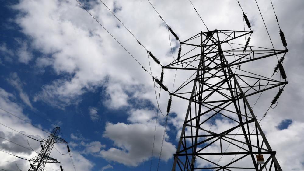 A pylon, viewed from the ground, with a second pylon to the left of the image. The sky is blue, but cloudy. The lattice framework of the pylon, its ceramic insulators and the wires can clearly be seen.