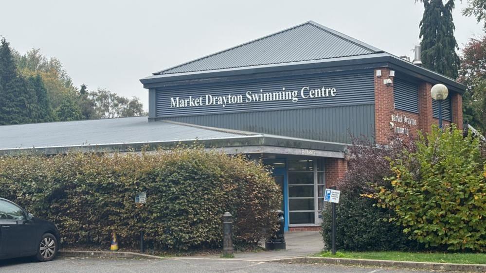 A photo of a grey building behind large green bushes. The building has large white letters on which read Market Drayton Swimming Centre.