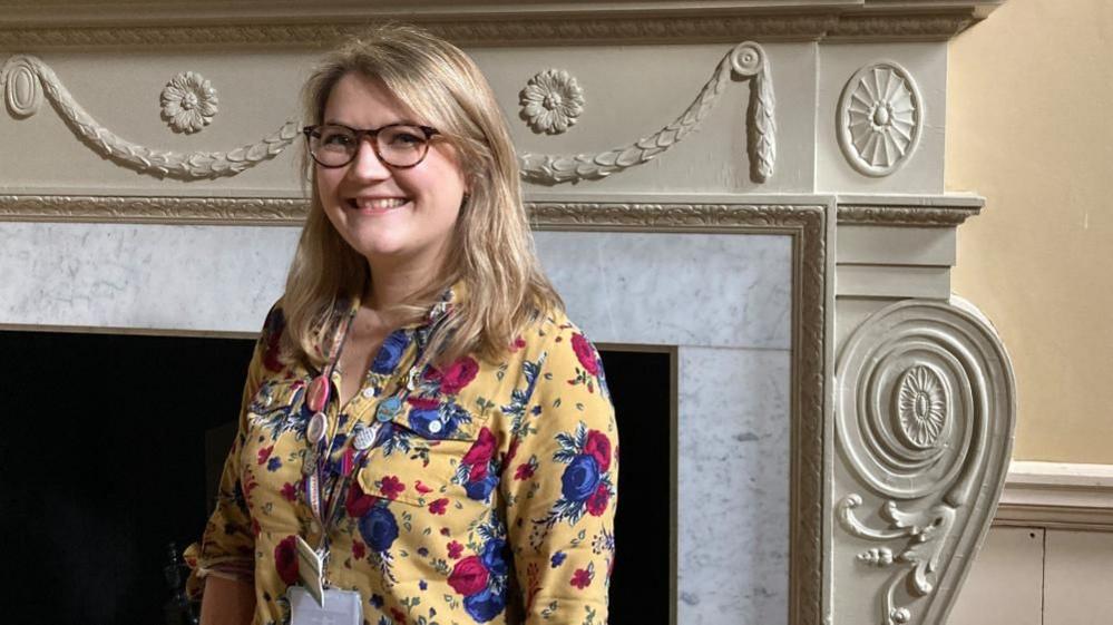 A woman with long blonde hair and glasses, wearing a yellow long sleeved shirt with red and blue flowers standing in front of a grand marble fireplace.