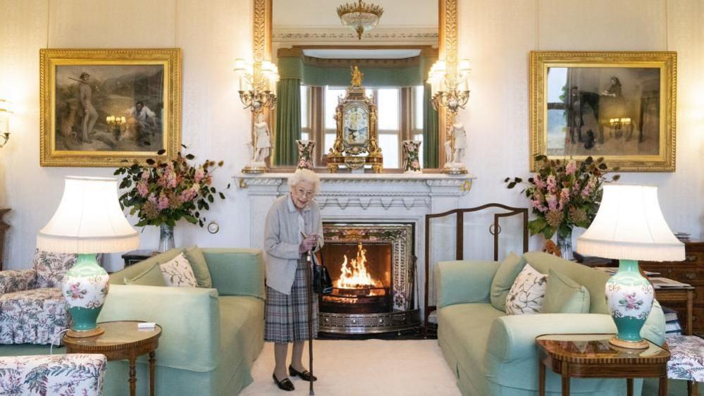 Queen Elizabeth II standing in the drawing room at Balmoral. The room is decorated with green and pink florals. There is a fireplace in the centre of the wall with a large mirror above it. Two paintings hang either side of the mirror. 