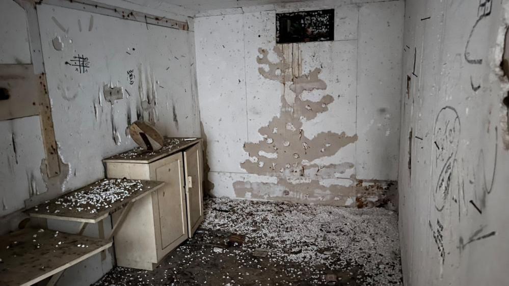 The bunker interior, with deteriorated polystyrene insulation on the walls, with graffiti, debris on the floor and a cupboard and shelves on the left and a ventilation shaft central on the upper part of the back wall