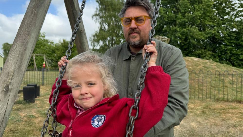 Matthew Lawson has greying hair, and sports a full beard and moustache. He wears brown-framed glasses, and is holding onto his daughter who is sitting on a swing in Cantley Village Hall playground. She has blonde hair and is wearing a red fleece with the school logo embroidered onto it. She is holding onto the chains of the swing. Behind them is other play equipment, fencing, a mound covered in grass and some trees.