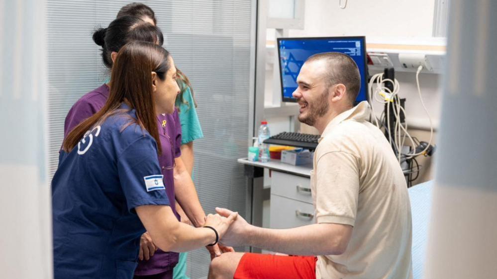 Smiling, Andrey Kozlov holds hands with woman whilst sat on hospital bed