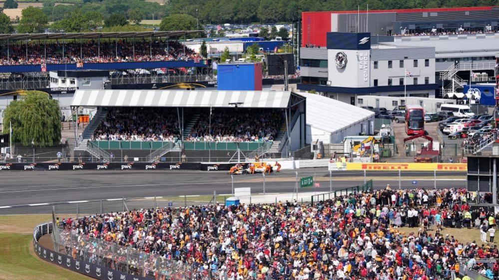 Silverstone racetrack showing a race in action, lots of crows in stands around the track and in viewing areas. There are stands, vehicles, and trees in the distance.