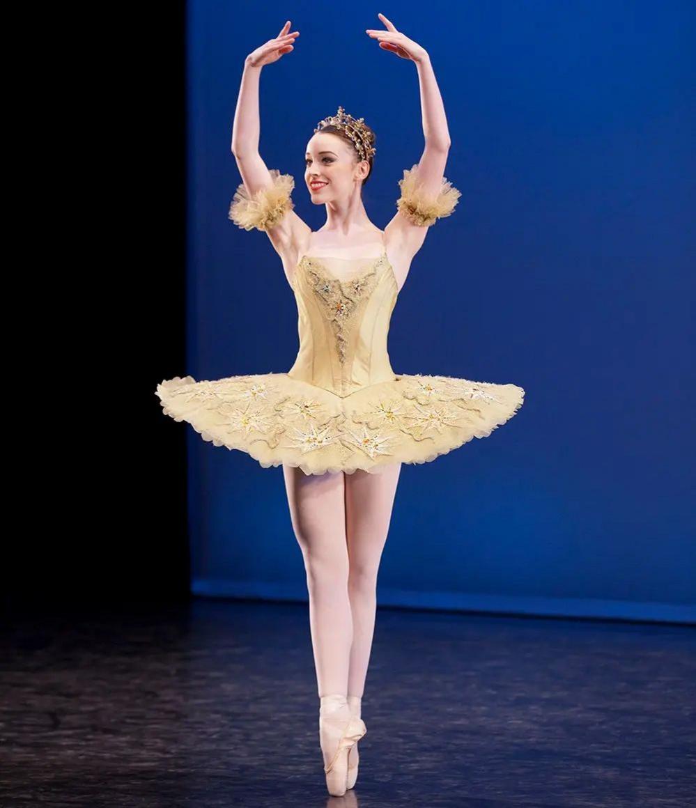 Ellen Elphick, smiling, in a cream tutu wearing a crown. She is en pointe and her arms are elegantly extended above her head. There is a royal blue background behind her.   