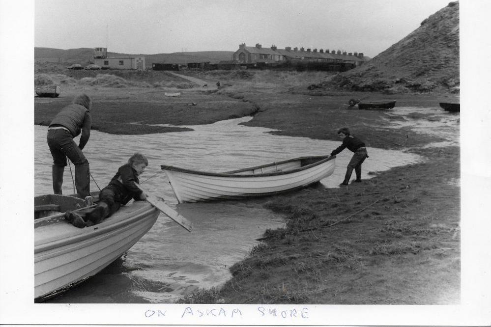 A black-and-white beach scene of three boys playing on two white row boats in the sea. Terraced houses are in the background.