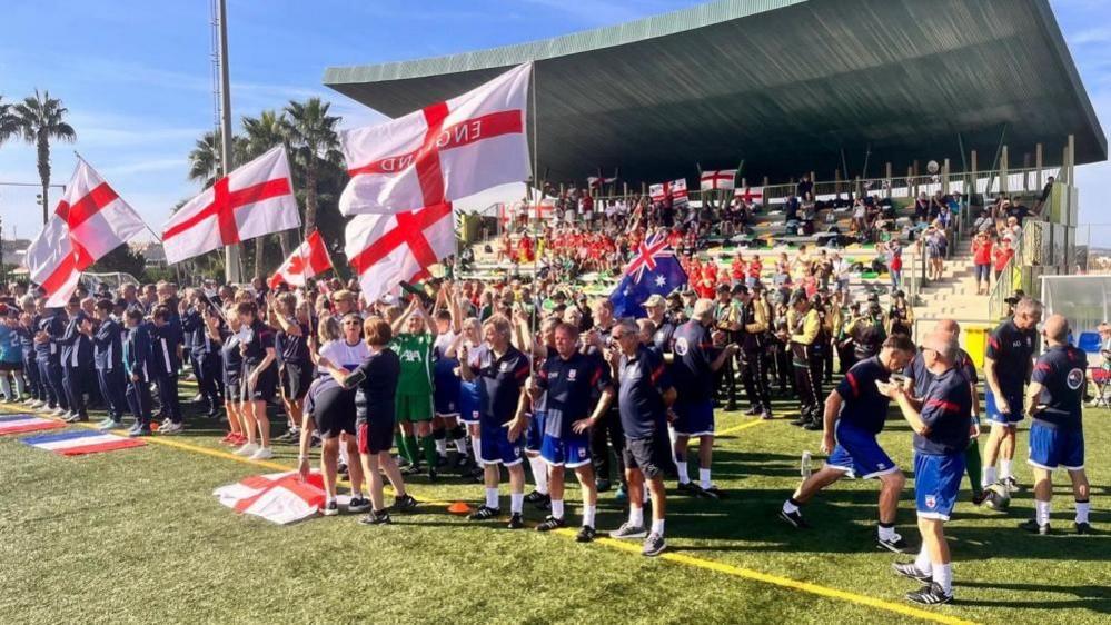 A crowd of people, England players and supporters, stand by the side of the pitch, waving England flags.