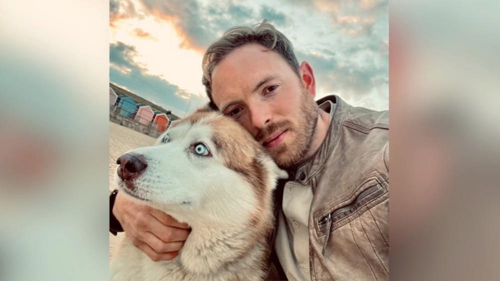 Ben Gladman, wearing a light brown jacket, pictured with his husky/malamute dog called Dobby. He is on a beach and there are beach huts behind him.