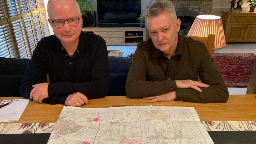 Two men looking at a map whilst sat at a table in a house. The lounge area can be seen behind them. One wears glasses and has short hair and wears a black shirt. The other has grey hair and a brown/green shirt.
