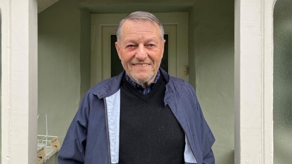 Paul Kelly stands in the porch doorway of his house. He wears a blue waterproof jacket, black jumper and blue checked shirt.