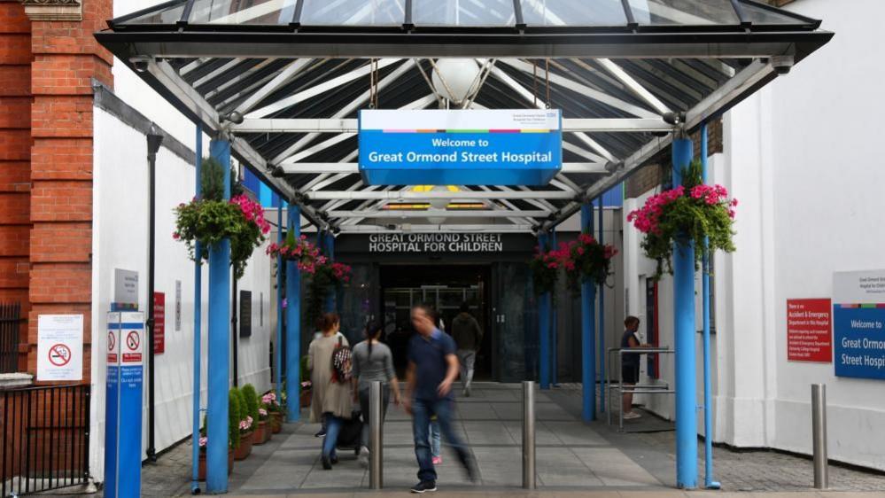 The entrance of Great Ormond Street Hospital, with a blue NHS sign above the walkway reading 'Welcome to Great Ormond Street Hospital' underneath a small covered area. Further back, there is a black sign above the main doors which reads in white writing, 'Great Ormond Street Hospital for Children'.