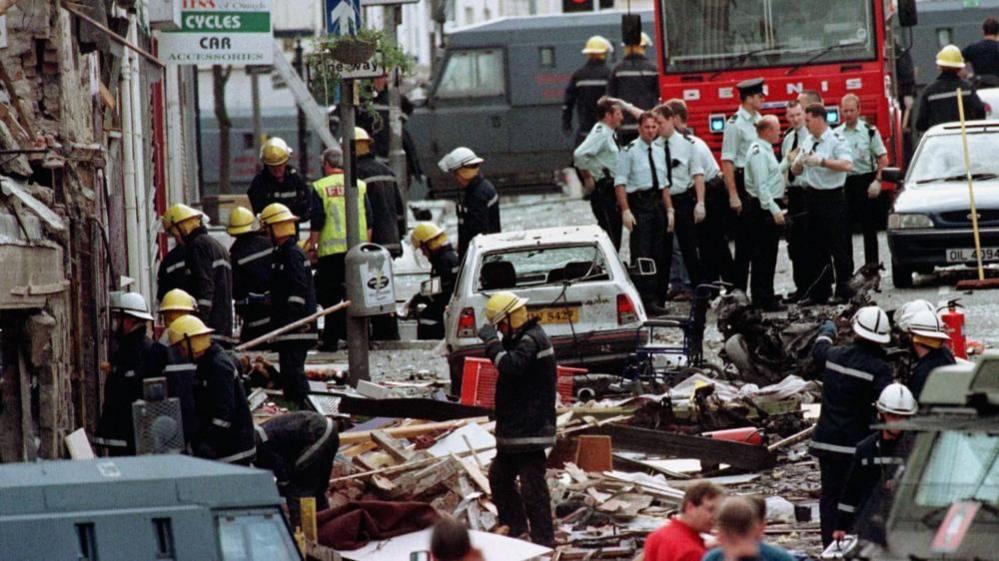 An image of emergency services including police officers, fire men and ambulance staff searching rubble along the main street of Omagh.
