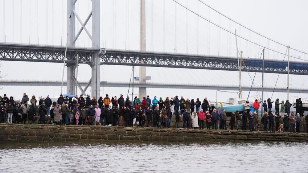 Spectators wrapped up in warm clothing watch the event on a stone pathway in front of the Forth Road Bridge.