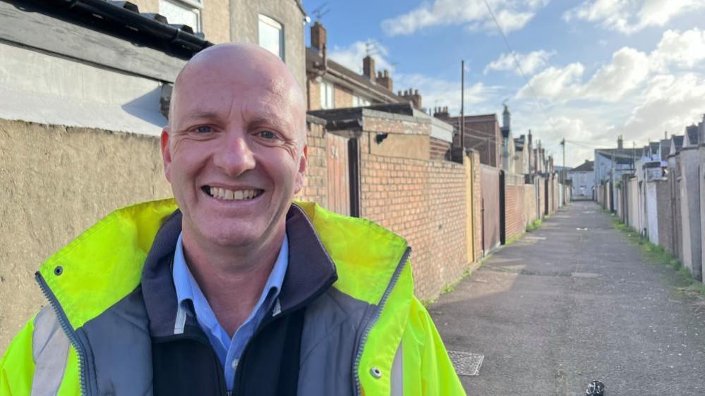 James Wilson, wearing a high visibility jacket and black fleece and blue shirt. He is smiling and standing in an alley cleared of bins and fly-tipped rubbish.