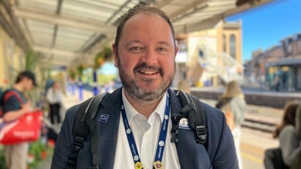Alex Hornby smiles at the camera, wearing a Northern lanyard and rucksack, standing on the platform at Harrogate Railway Station.