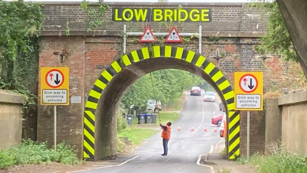 A man in an orange hi-vis jacket holds a stick which he is using to touch the underside of a bridge