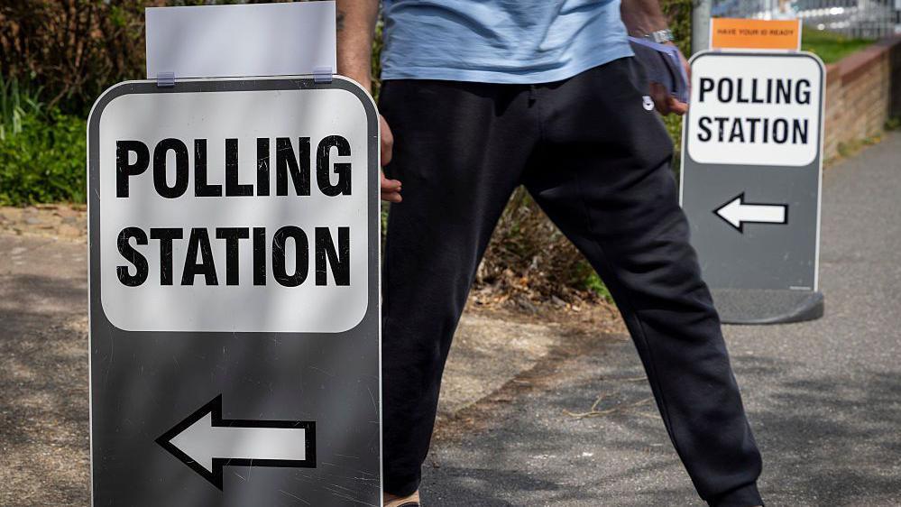 A person in black trousers leaves a polling station. He walks past a sign saying polling station with an arrow.
