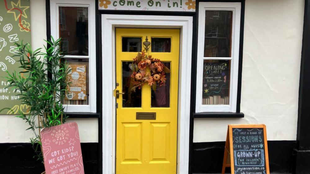 The photo shows a bright yellow door decorated with an autumn wreath. Above the door is a sign to say Come on in. Two chalkboard signs advertising sessions and activities are either side of the door.  