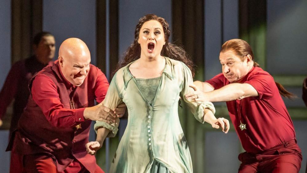 Claire Lees playing Pamina during a dress rehearsal of Opera North's production of Mozart's The Magic Flute, at Leeds Grand Theatre. She is wearing a green dress and is open mouthed, with two men dressed in red shirts and trousers, one either side, holding on to her arms in a pulling motion.