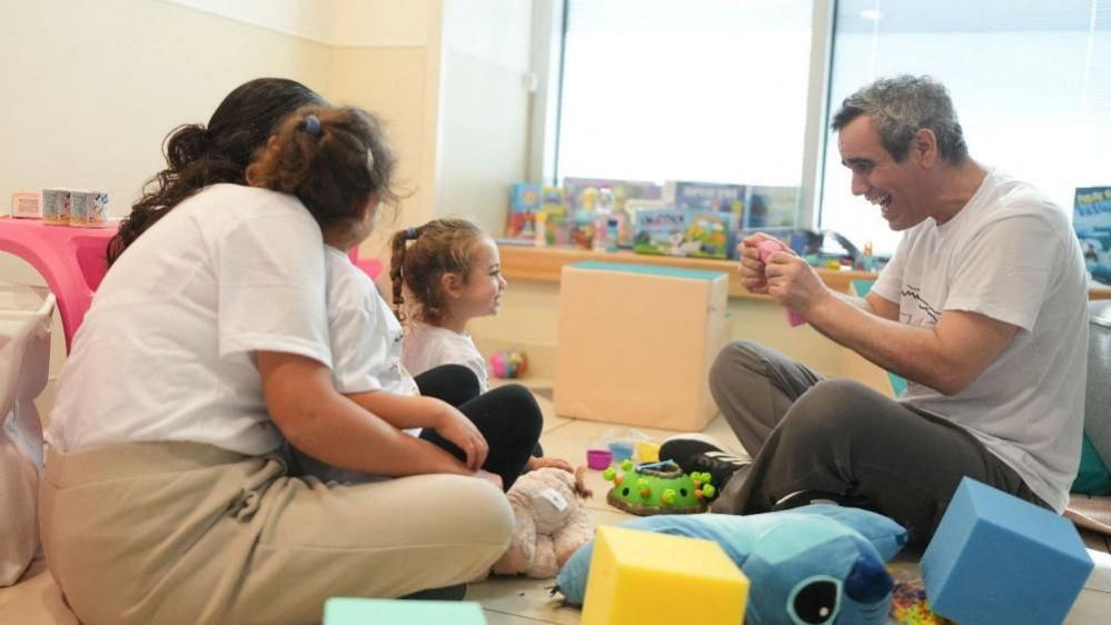 Omri Miran, wearing a white T-shirt and grey trousers, salt-pepper hair, is seen sitting in a room with children's toys, smiles as he looks at his daughter, also sitting and smiling, with two other women watching her