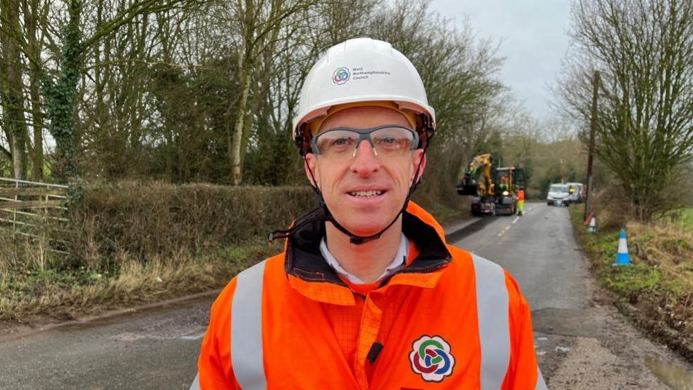 Peter Ingram wearing a white hat and glasses looking at the camera with orange hi-vis. A road lined with trees is behind him, and there is a yellow digger in the background