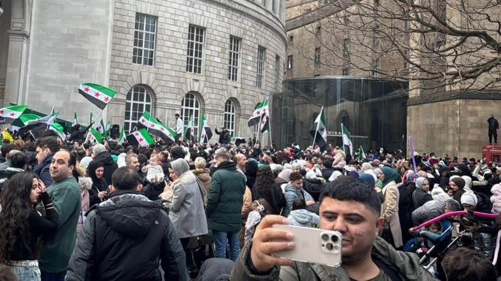 A large crowd of people stand in front of Manchester Town Hall. Many people are waving Syrian flags