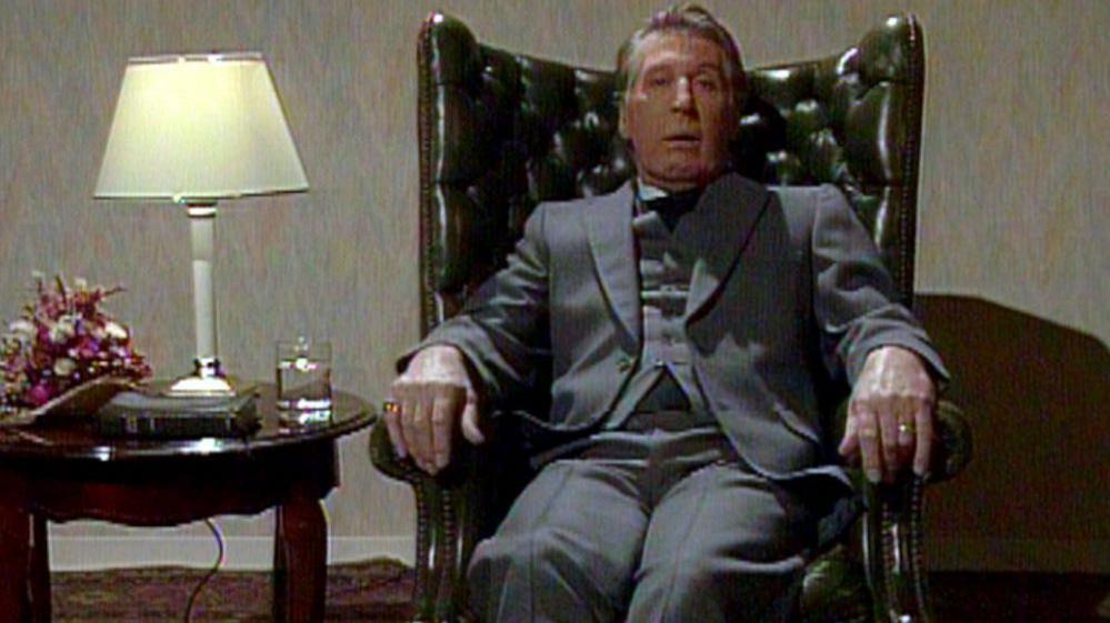 Rikki Fulton in character as Rev IM Jolly. He is sitting in a green, leather  armchair, wearing a grey suit and dog collar. Next to him is a table with a lamp, flowers and glass on it.