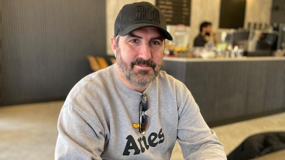 A man sitting in a coffee shop, with a cap on his head, wearing a grey jumper and glasses round his neck. The coffee bar is blurred behind him.