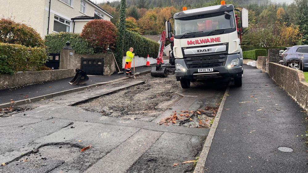 A large lorry mounts a damaged road. A worker is beside a digger with a shovel. The road has large chunks of tarmac out of it. 