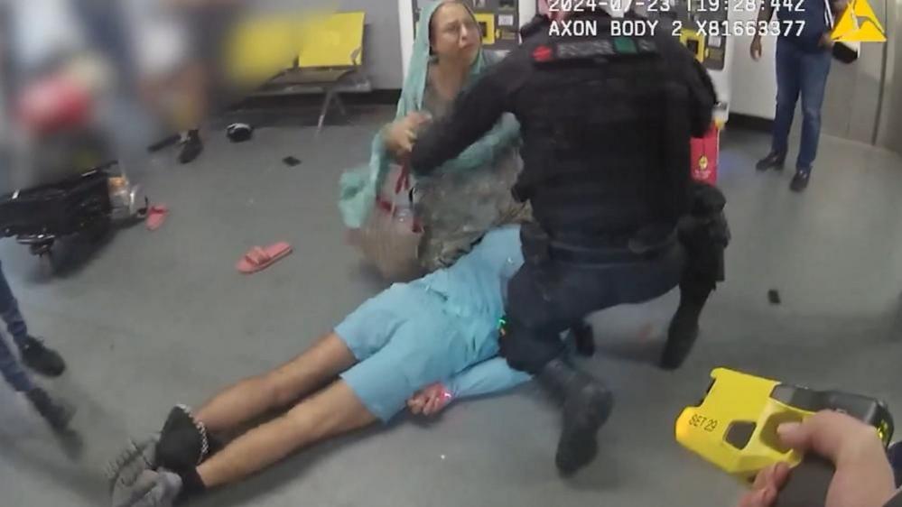 A screenshot from police bodyworn footage of a man lying on the floor on his front. A male police officer and a woman are kneeled over him. In the foreground, someone is holding a Taser device.