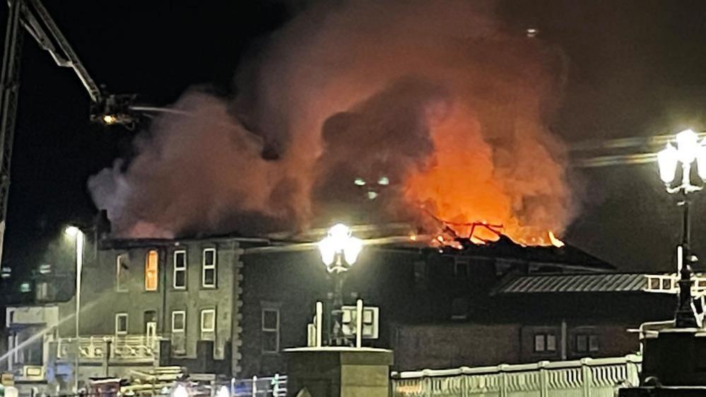 The Haven Bridge pub at night, with its roof lost and flames lighting up the sky, and smoke emitting from the top of the building. Fire appliances, including an aerial ladder platform are visible on both sides of the image, and the Haven Bridge, with illuminated street lights bleaching spots on the image, over the River Yare is visible in the foreground.