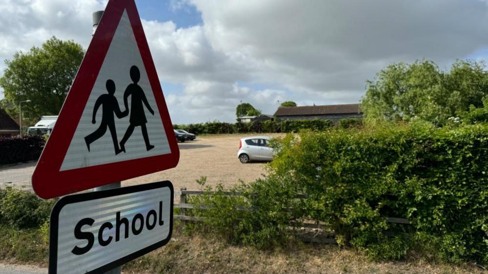 A close up shot of a road sign bearing a parent and child emblem, and the word "School" on a separate sign beneath. In the distance are wooden buildings that form part of Cantley Primary School. In the foreground is a car park with a few cars parked, and some hedging and trees. The sky is cloudy.