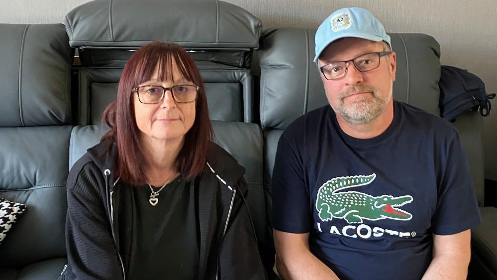 Louise Slater in a black hoodie and black t-shirt sits on a grey sofa next to Clint Slater who wears a blue t-shirt, glasses and a Coventry City FC cap.
