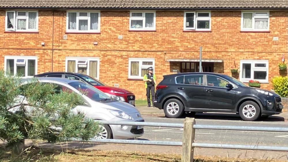 A police officer standing outside by a long row of brick houses, by a black car, with a silver car and a red car close by