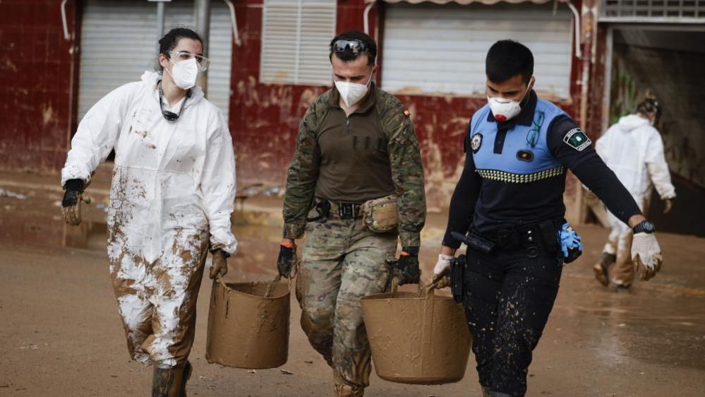 Three people wearing face masks and mud-splattered clothing carry buckets as they continue with cleaning operations in the flood-hit municipality of Paiporta