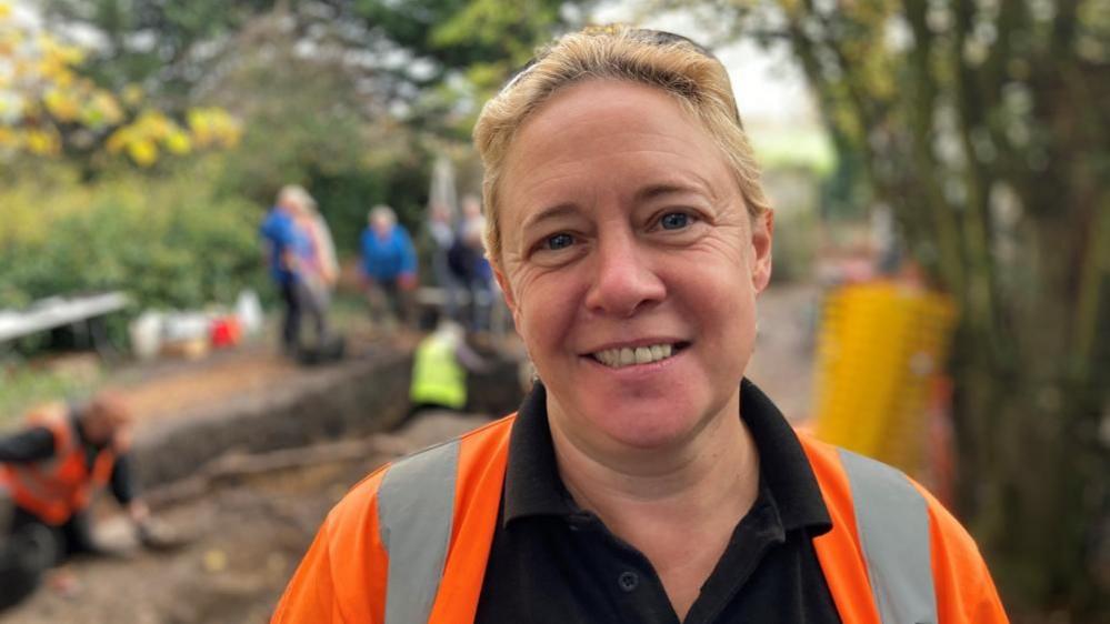 Dr Emma Watson has short blonde hair and is smiling. She is wearing a black polo short and a high-vis orange jacket. In the background, people are digging in a trench in the ground.
