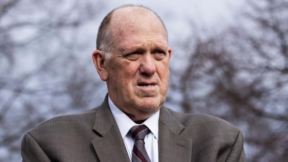 Tom Homan stands outside in front of leafless trees wearing a grey suit.