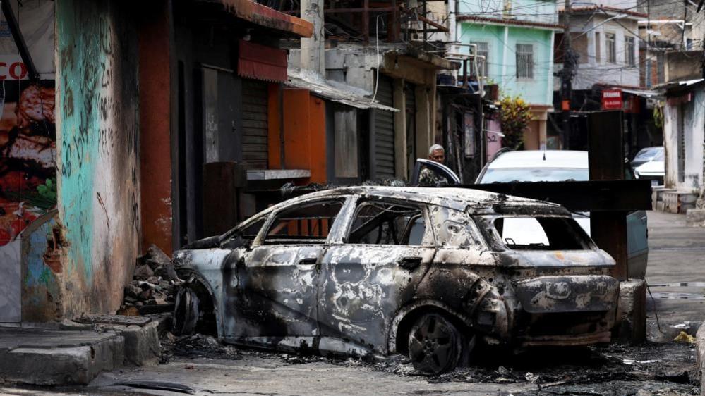 A person looks on behind a burnt car during a police operation against drug trafficking at the favela do Penha, in Rio de Janeiro