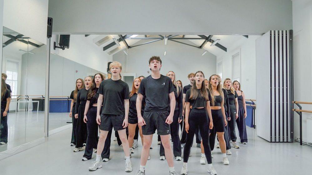 A group of young people standing in a group singing. They are all wearing dark clothing and standing in a white rehearsal room with mirrors on the wall to the left.