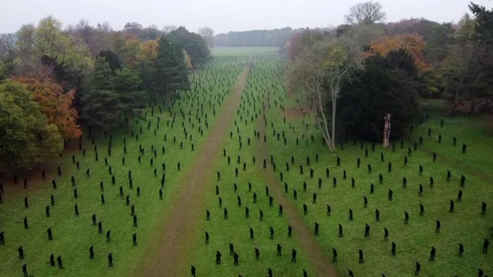 An aerial photo of thousands of silhouettes of servicemen in Stowe Gardens. The figures are black and scattered across green grass with trees either side.