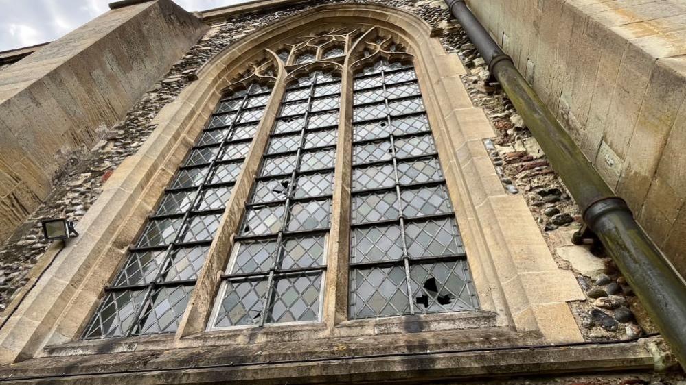 One of the large north-facing windows on St Nicholas Minster; a trio of gothic arched window apertures, with diamond leaded glass set within, featuring a rectangular border. The window is made of sandstone and set within a flint wall.