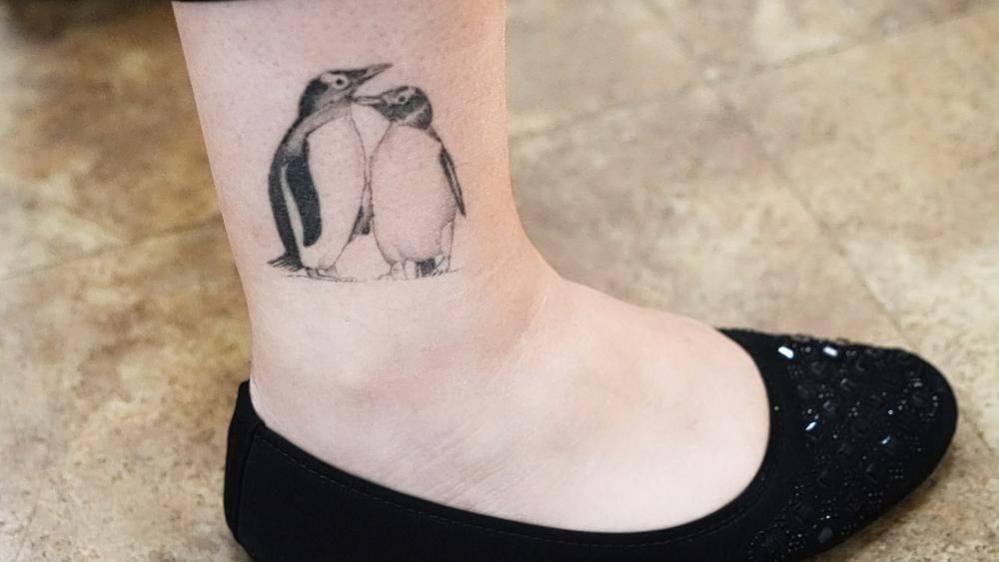 Tattoo of two penguins on a woman's lower leg. She is wearing a black shoe.