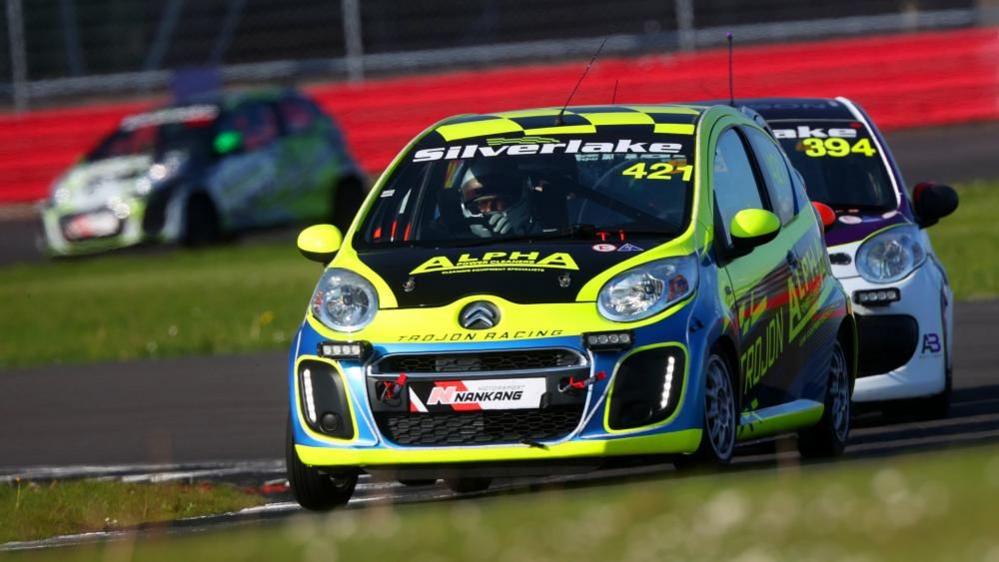 Yellow and white Citroen C1s racing around a circuit. Both have Silverlink banners with white writing across the top of the windscreen. The front car is numbered 421 and the rear car is 394. The front also has "Alpha" under the windscreen. Another Citroen C1 is in the background in front of a red barrier.