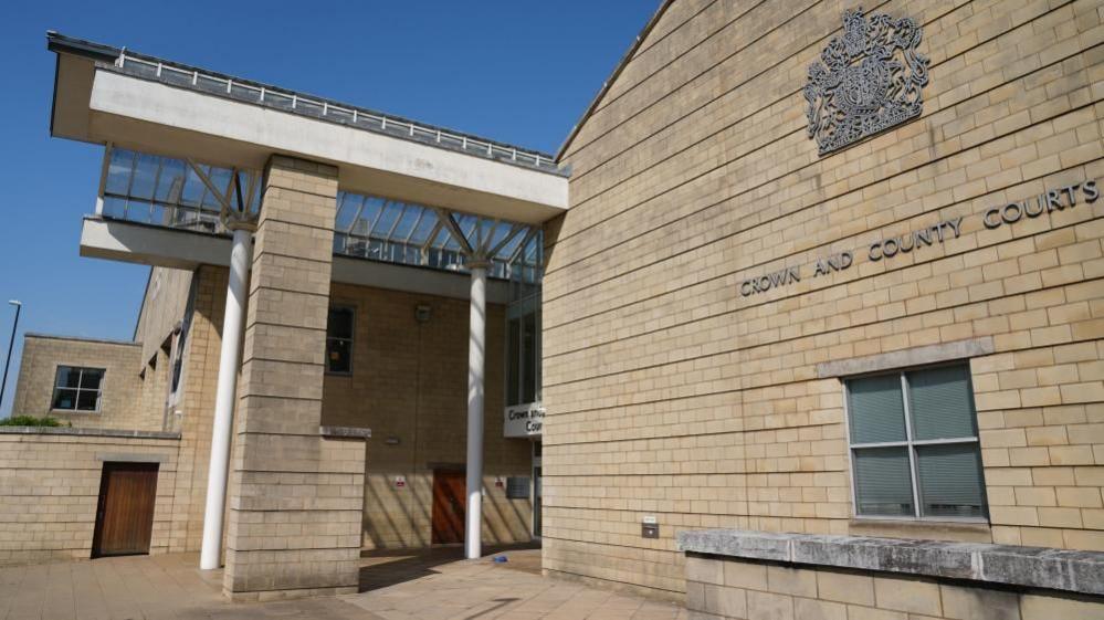 Court building in light brown brick with glass-covered entrance supported by white pillars. "Crown And County Court" is visible on a wall to the right.