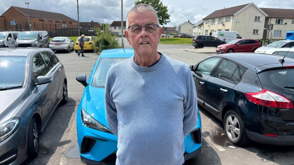 Llun o Steve Collins yn edrych at y camera. Mae e'n sefyll mewn maes parcio ar ystad y Gurnos ar ddiwrnod braf.