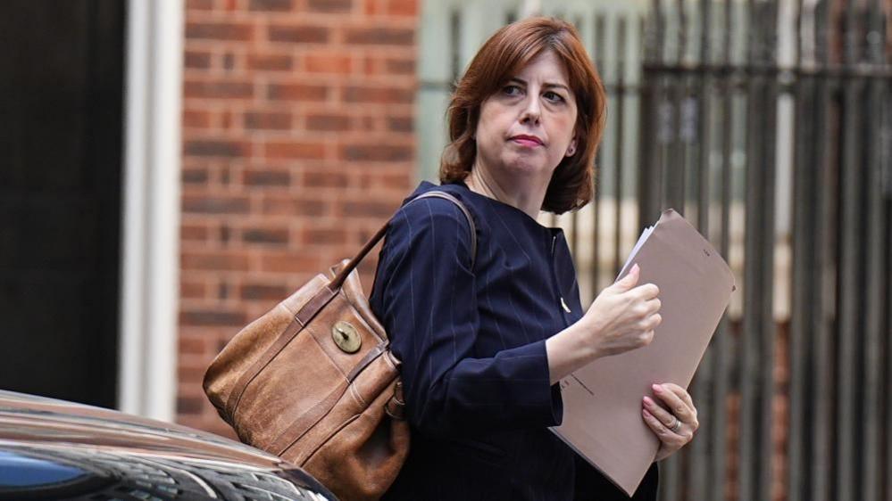 Lucy Powell is holding a brown folder and has a large brown-coloured bag over her right shoulder. She is wearing a dark blue jacket. A brick wall and a metal fence can be seen in the background.