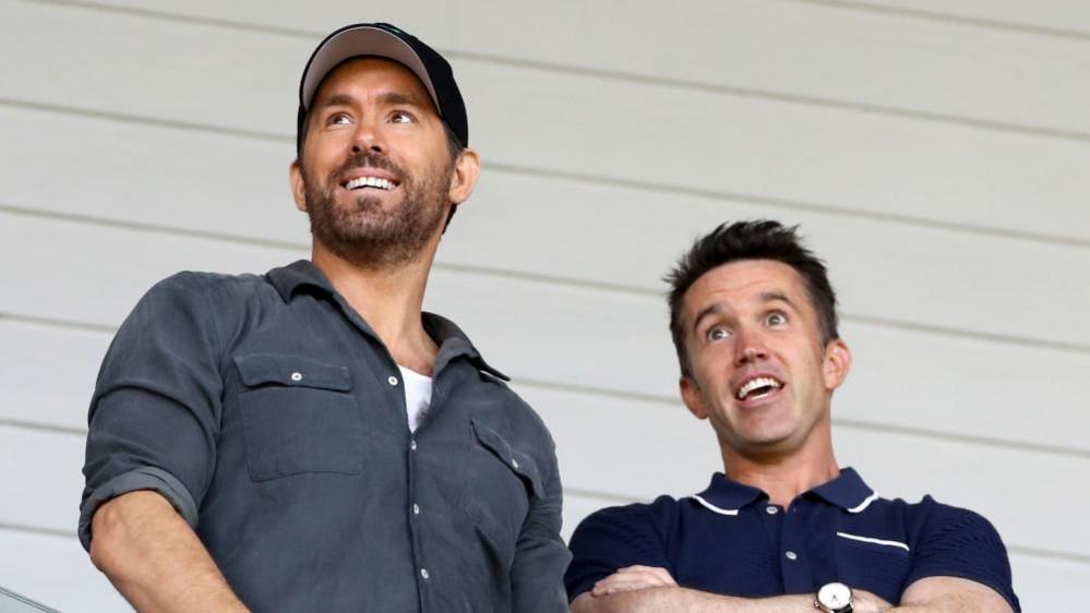 Ryan Reynolds in a cap and grey shirt stood alongside Rob McElhenney in a blue polo shirt. Both are smiling and appearing to watch a football match