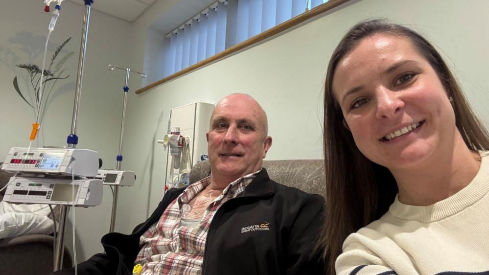 Angharad Binder, with long straight hair, wearing a white top with black stripes. She is next to her father Robert Bendall, who is undergoing treatment for bowel cancer. He is wearing a checked shirt and black jacket, and he has close cropped hair.