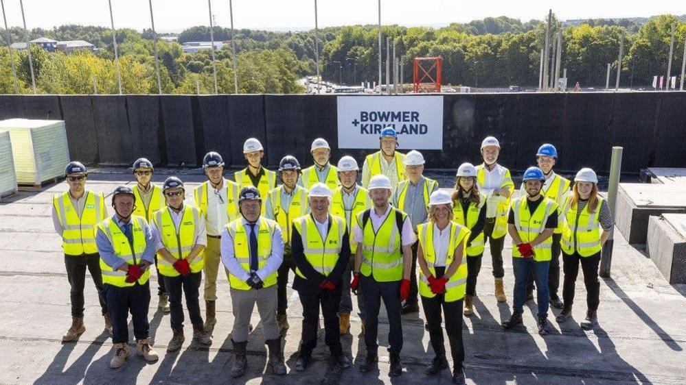 A group of 20 men and woman are standing on the roof space of an apartment block, hearing white and blue helmets and yellow high-visibility jackets. There is a sign in the background that reads "Bowmer and Kirkland".